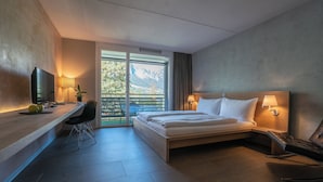 A hotel room with a bed, desk, chair, TV, and a view of mountains through a large window.