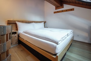 Una cama individual con estructura de madera y ropa de cama blanca en una habitación con suelo de madera y techo inclinado.