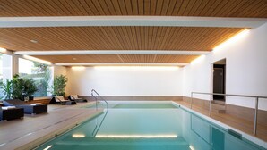 An indoor swimming pool with wooden ceiling, lounge chairs, and potted plants.