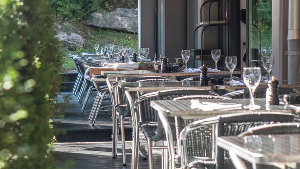 An outdoor dining area with tables and chairs, string lights, and a view of greenery.