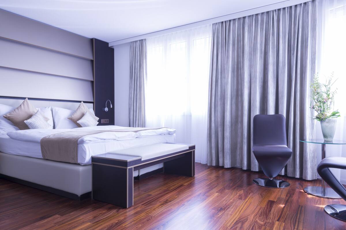 Ein modernes Hotelzimmer mit einem großen Bett, einem Nachttisch, einem Sessel und einem Fenster mit Vorhängen.