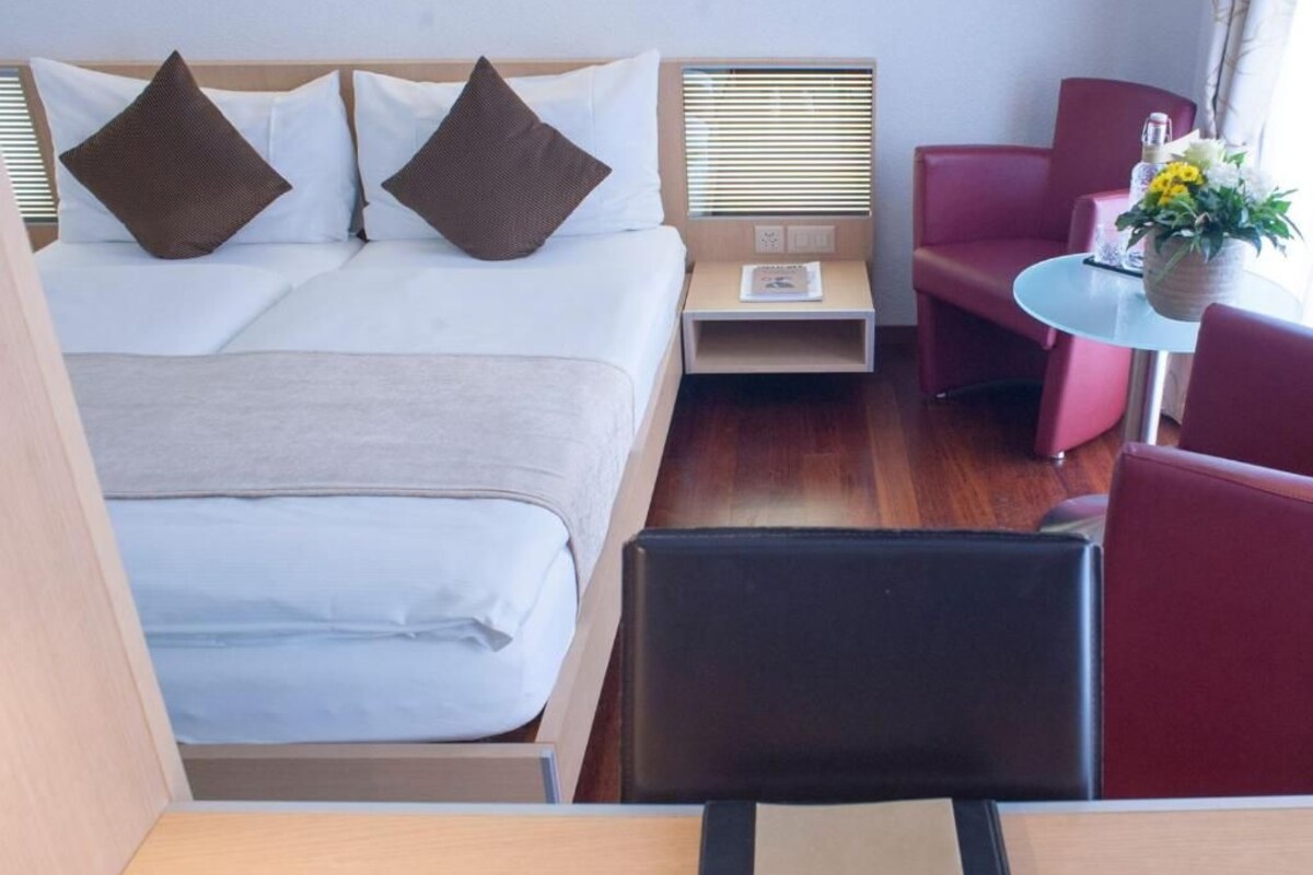 Habitación de hotel con cama, mesita de noche, una silla y una pequeña mesa con un jarrón de flores.