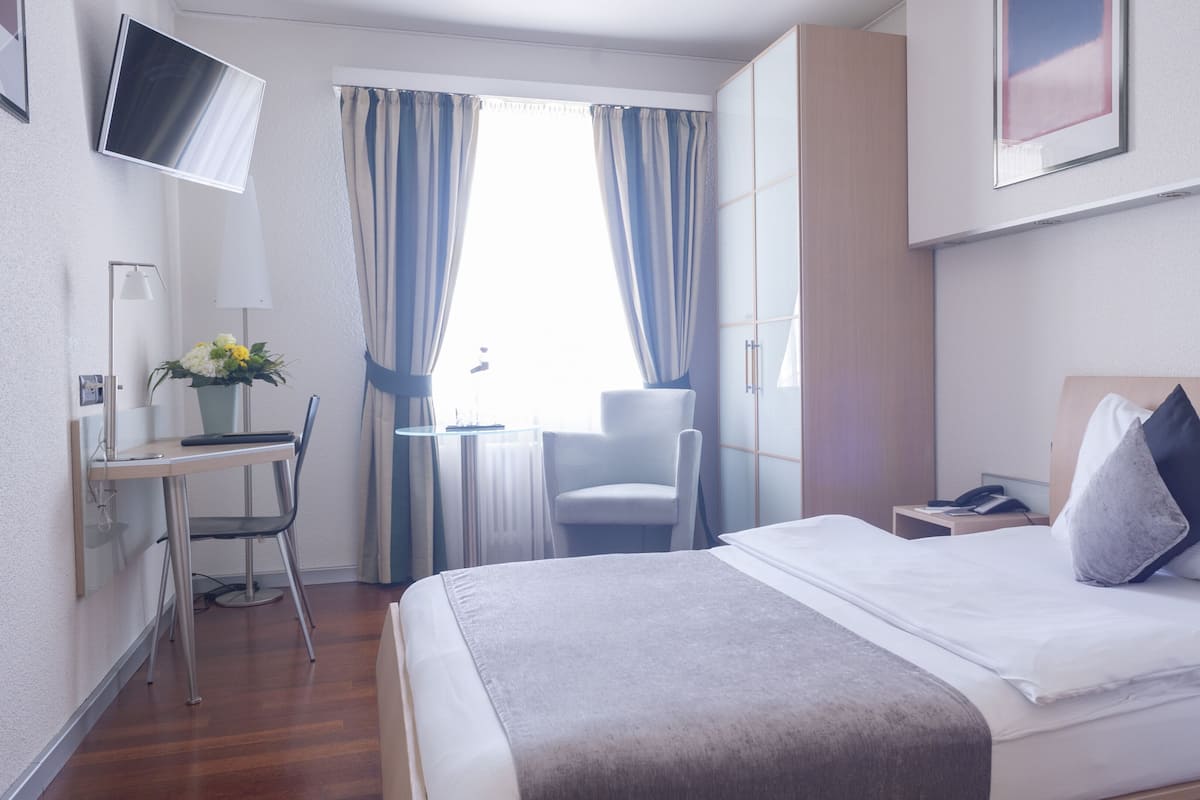 A hotel room with a bed, bedside table, chair, desk, TV, and curtains.