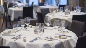 Un comedor con mesas redondas dispuestas para una comida, cada una con copas de vino, cubiertos y platos.