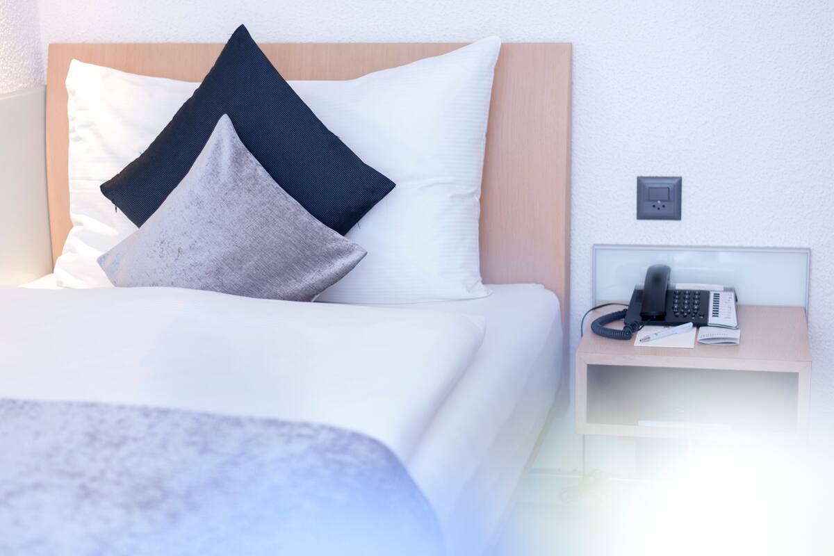 Ein ordentlich gemachtes Bett mit weißer Daunendecke und zwei Kissen, ein Nachttisch mit Telefon und Stift sowie eine Steckdose.