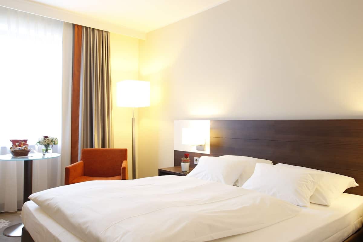 Une chambre d’hôtel avec un grand lit, une chaise, une petite table avec une lampe et une fenêtre avec des rideaux.