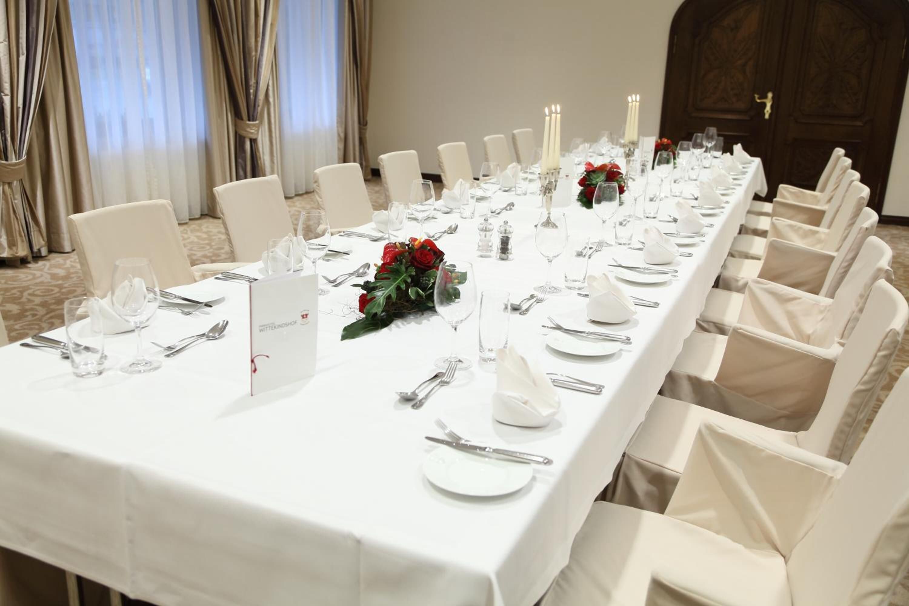 Ein langer Esstisch, festlich gedeckt für eine Veranstaltung, mit weißen Tischdecken, ordentlich drapierten Gedecken und floralen Mittelpunkten.
