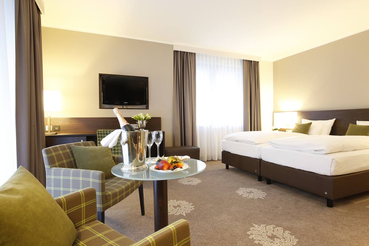Une chambre d’hôtel avec un lit, deux fauteuils, une petite table sur laquelle se trouve une assiette de fruits, une télévision et une fenêtre avec des rideaux.