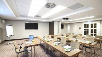 Un salón de conferencias con largas mesas, sillas, una pizarra y una pantalla de proyector.