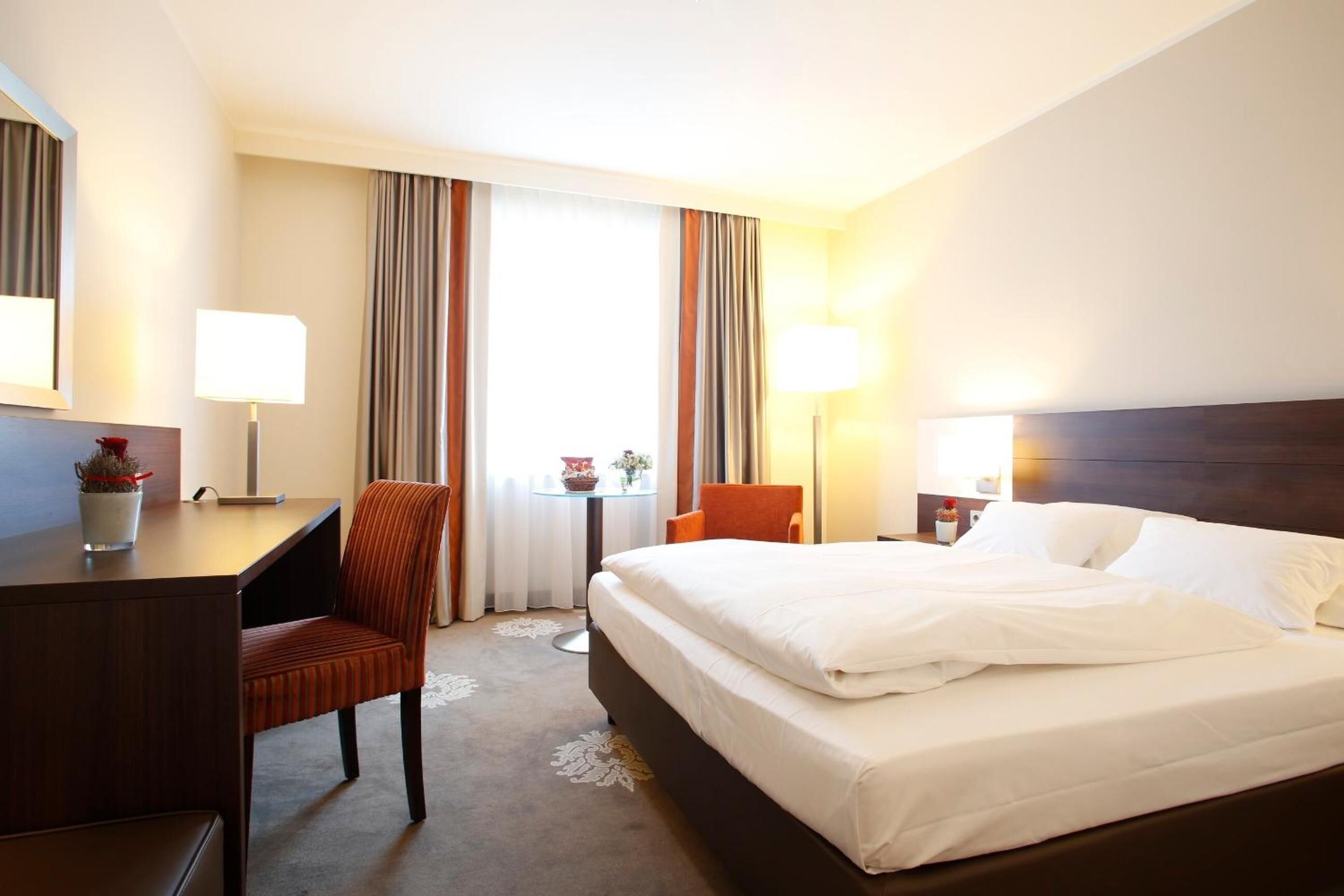 A hotel room with a bed, a desk, a chair, a lamp, and a window with curtains.