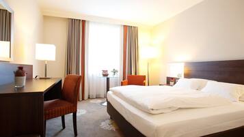 A hotel room with a bed, a desk, a chair, a lamp, and a window with curtains.