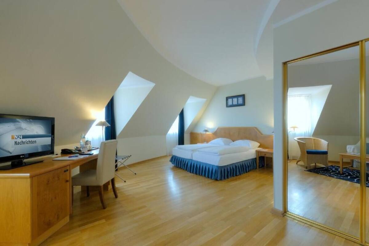 A modern hotel room with a wooden desk, a bed, a chair, and a TV.