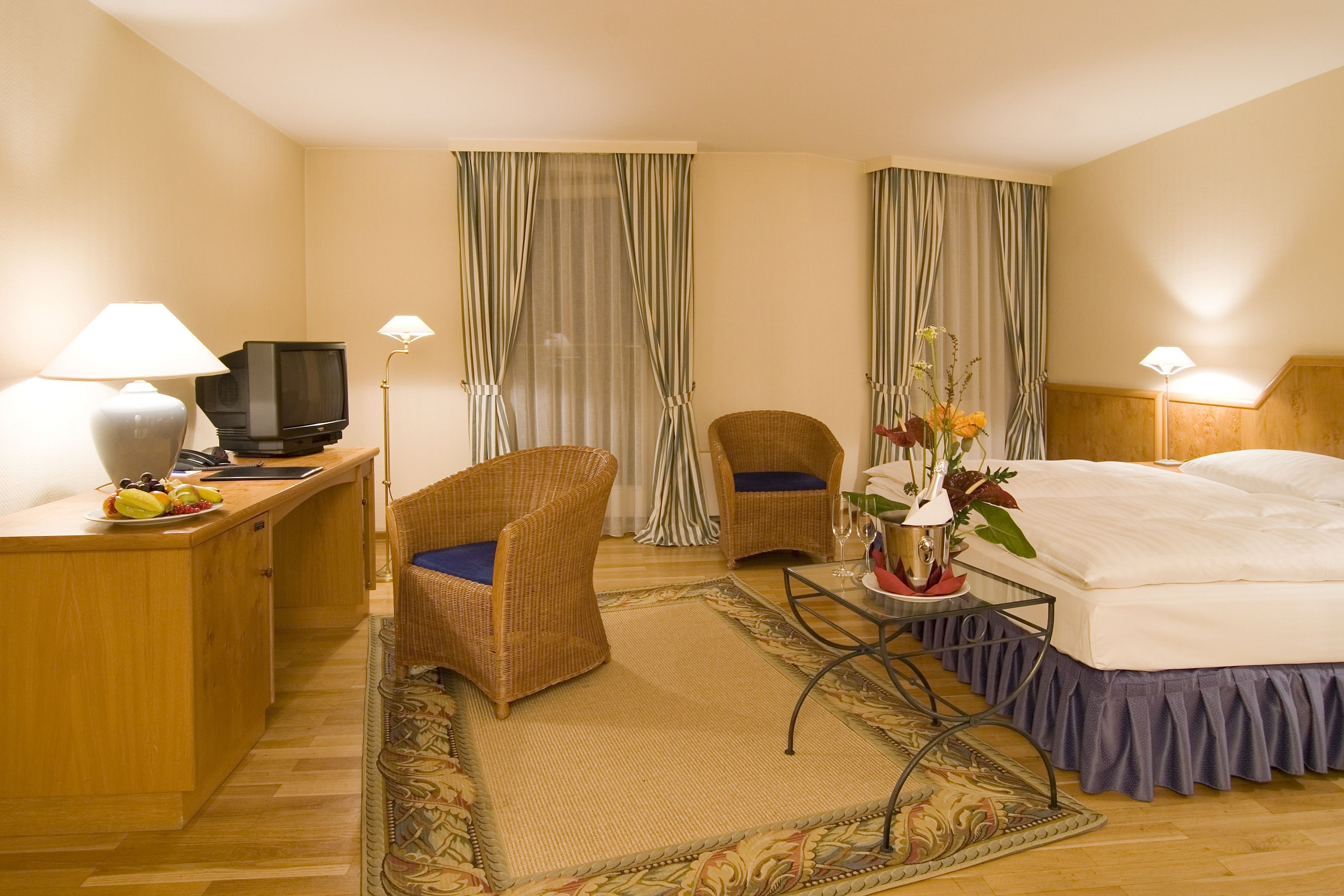 A hotel room with a bed, two chairs, a desk with a television, and a small table with a vase of flowers.