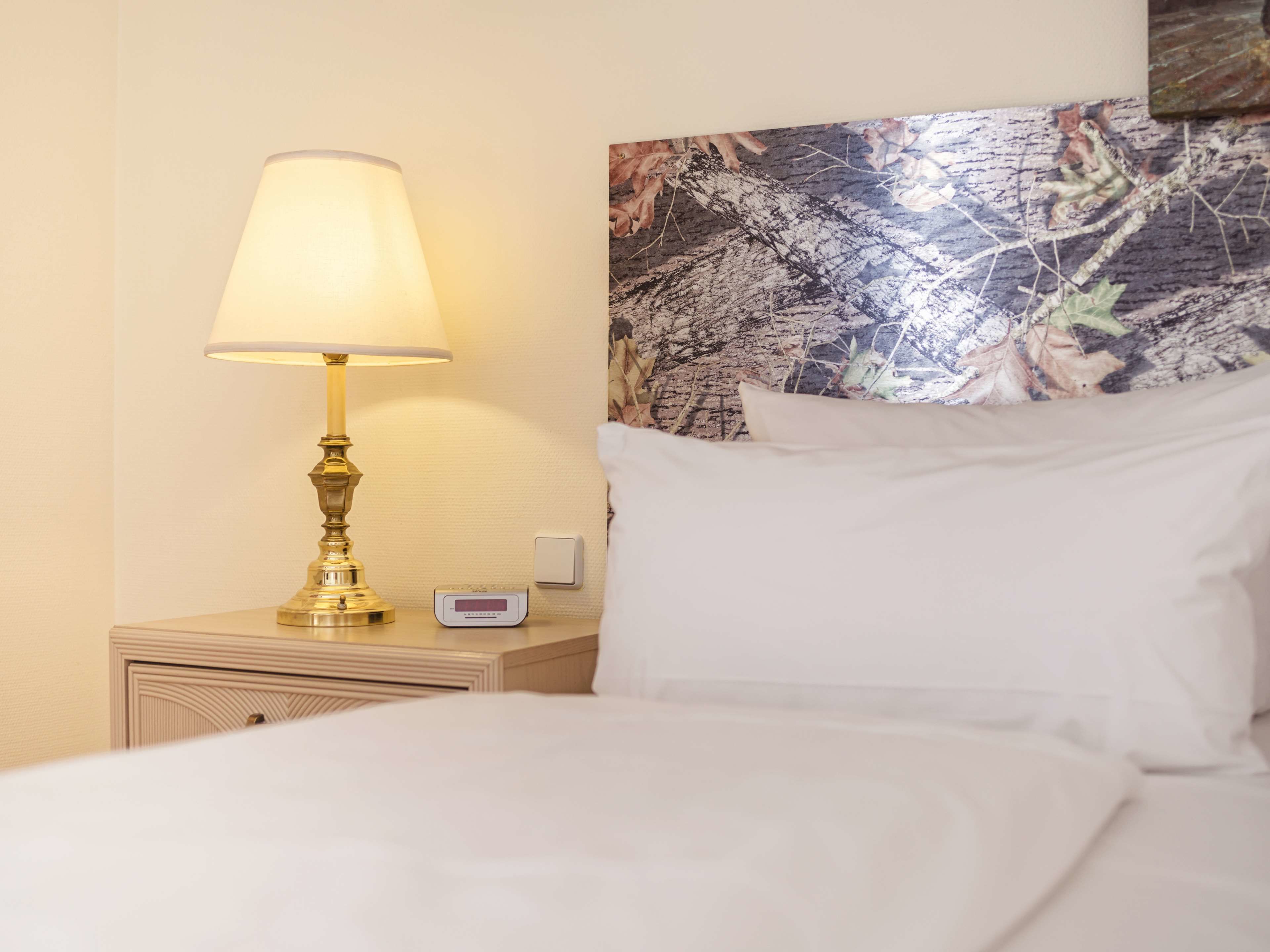 A neatly made bed with white linens, a bedside lamp, a digital thermostat, and a wall with a nature-themed artwork.