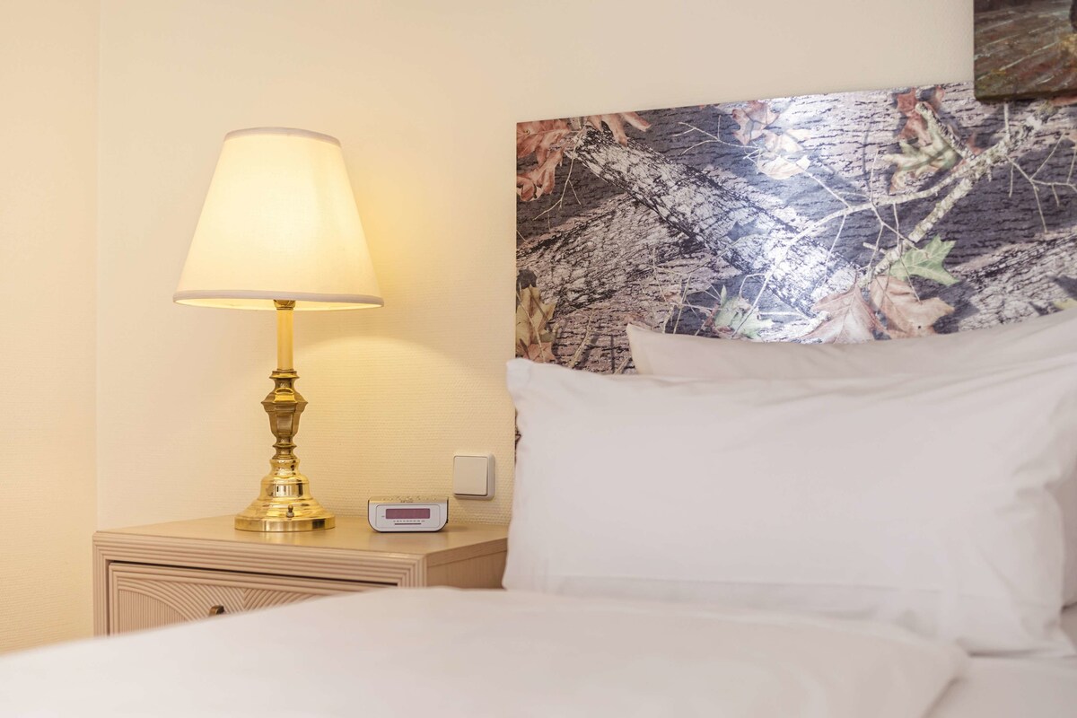 Un lit bien fait, avec du linge de lit blanc, une lampe de chevet, un thermostat numérique et un mur orné d’une œuvre d’art sur le thème de la nature.