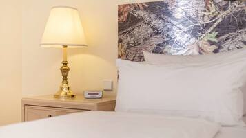 A neatly made bed with white linens, a bedside lamp, a digital thermostat, and a wall with a nature-themed artwork.