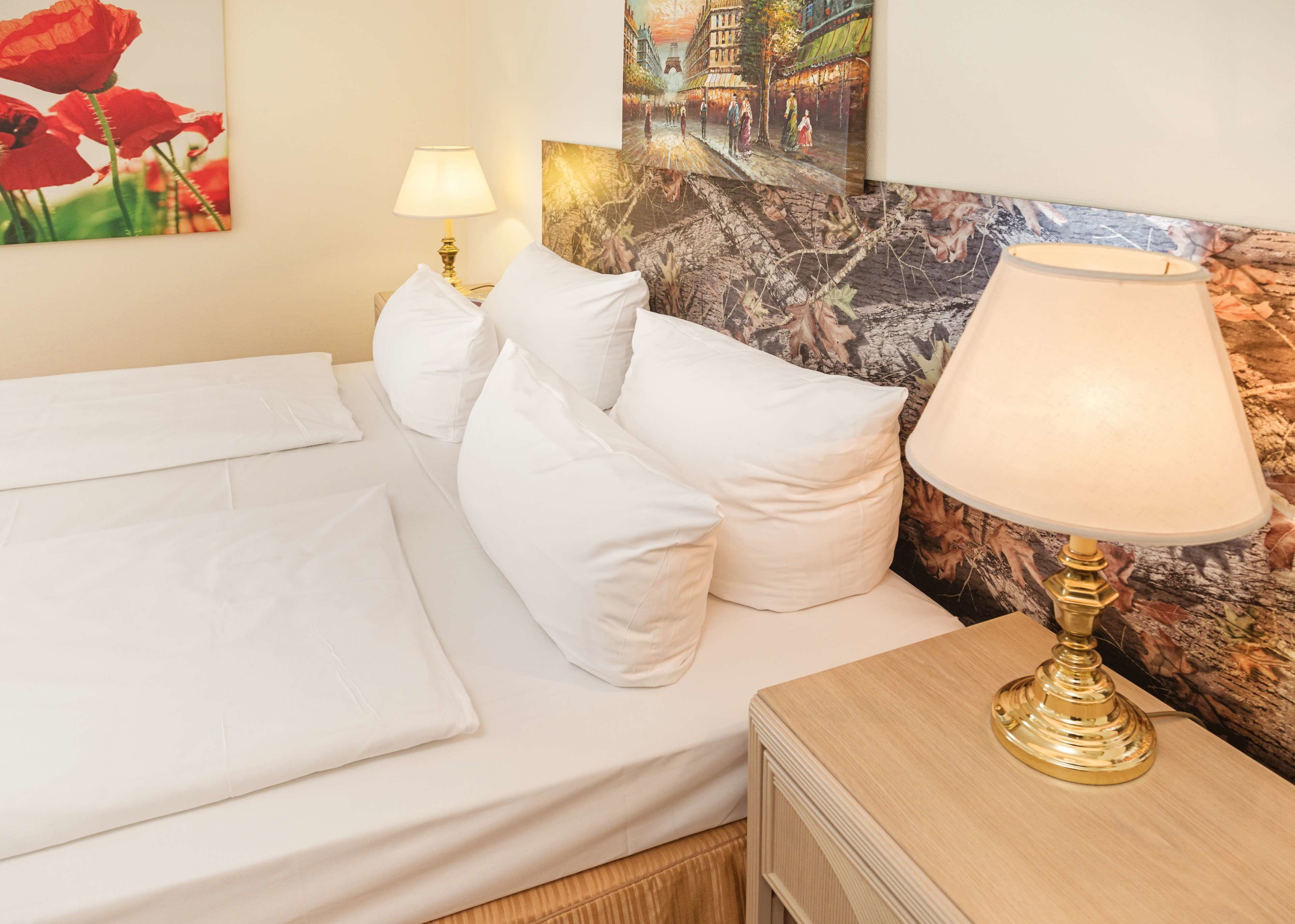 Een netjes opgemaakt bed met witte lakens en kussens, een nachtkastje met een lamp, en een muur met een schilderij.