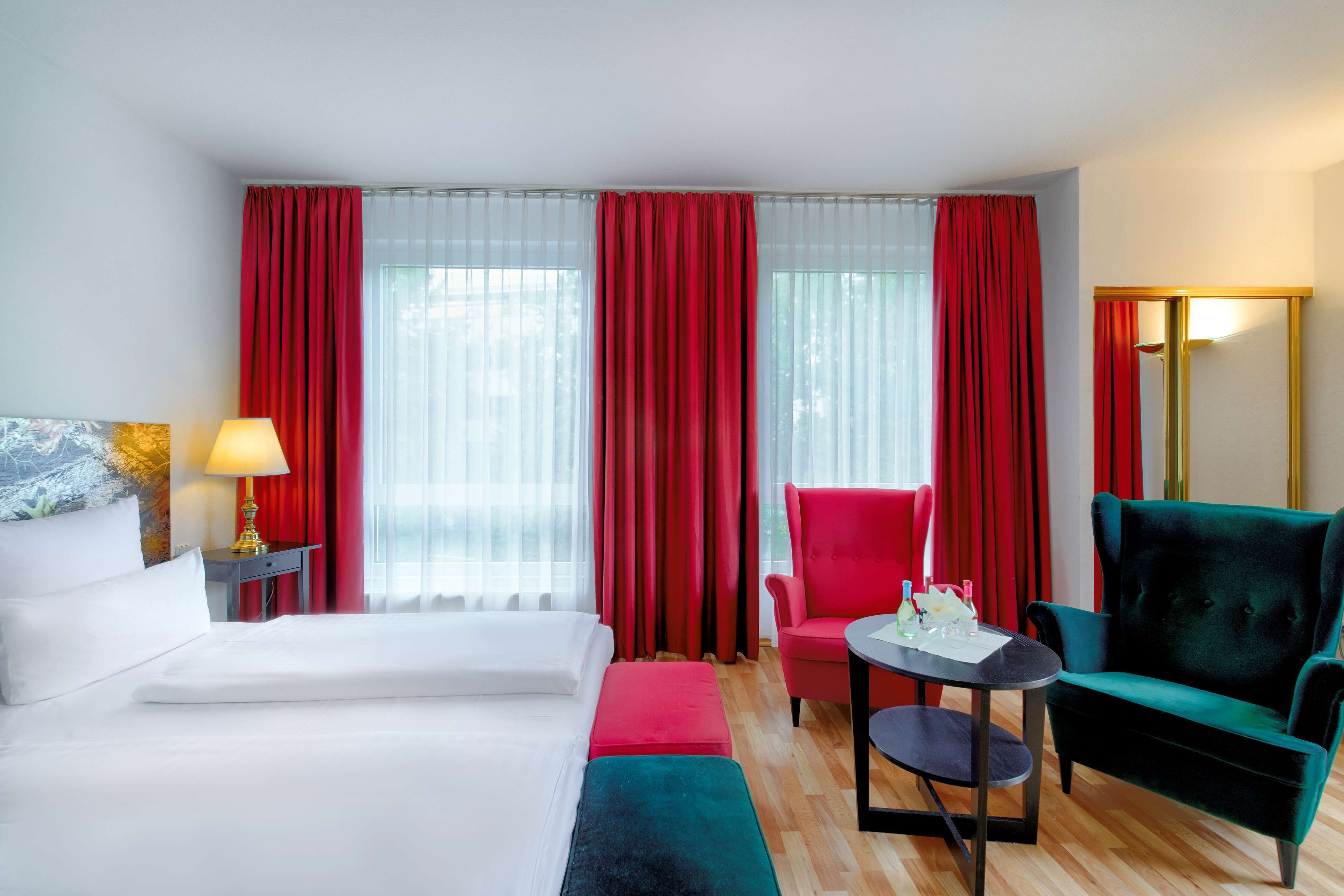 A hotel room with a bed, a red armchair, a green armchair, a small table, and a lamp.