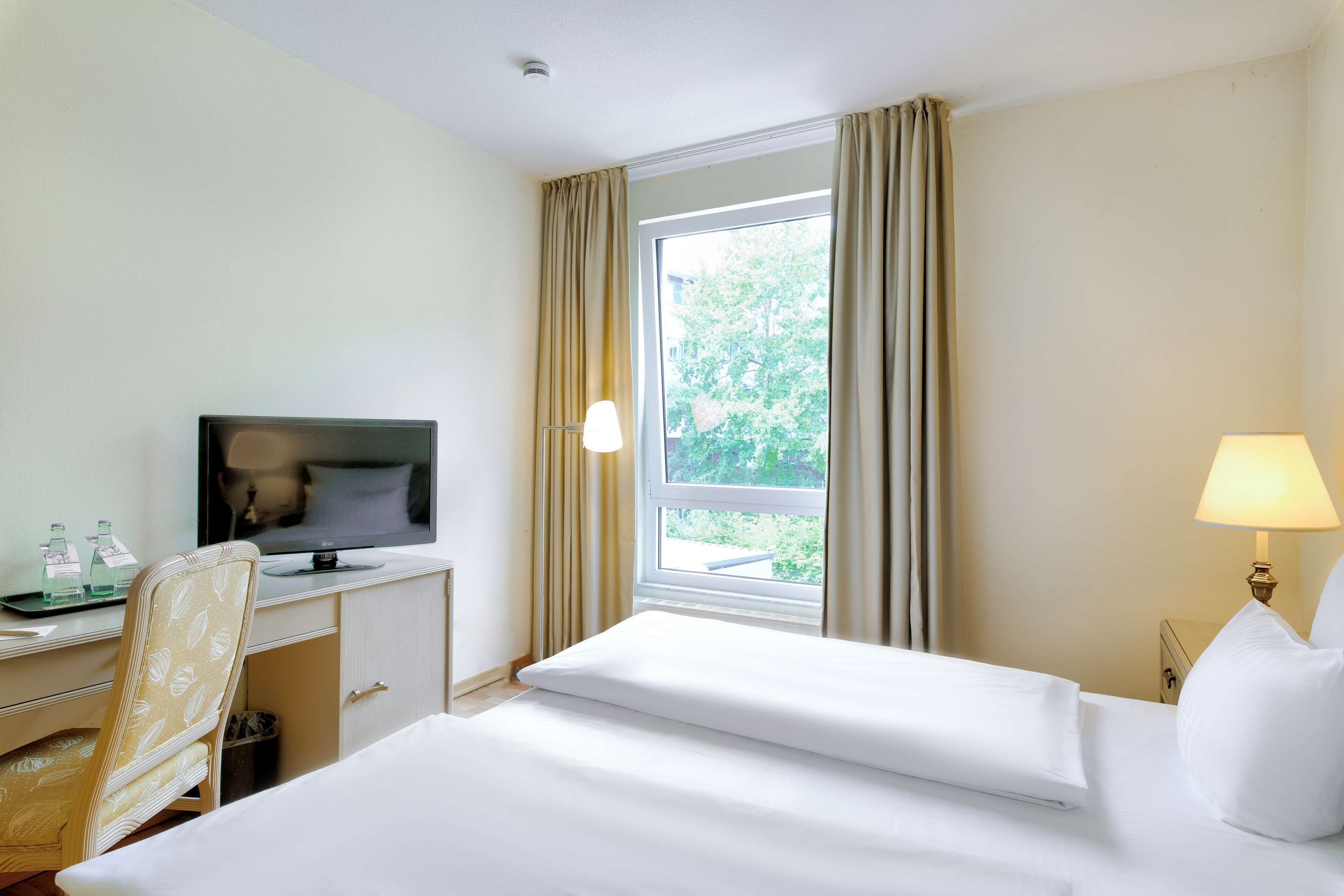A hotel room with a bed, a desk with a television, a chair, a window with curtains, and a bedside lamp.