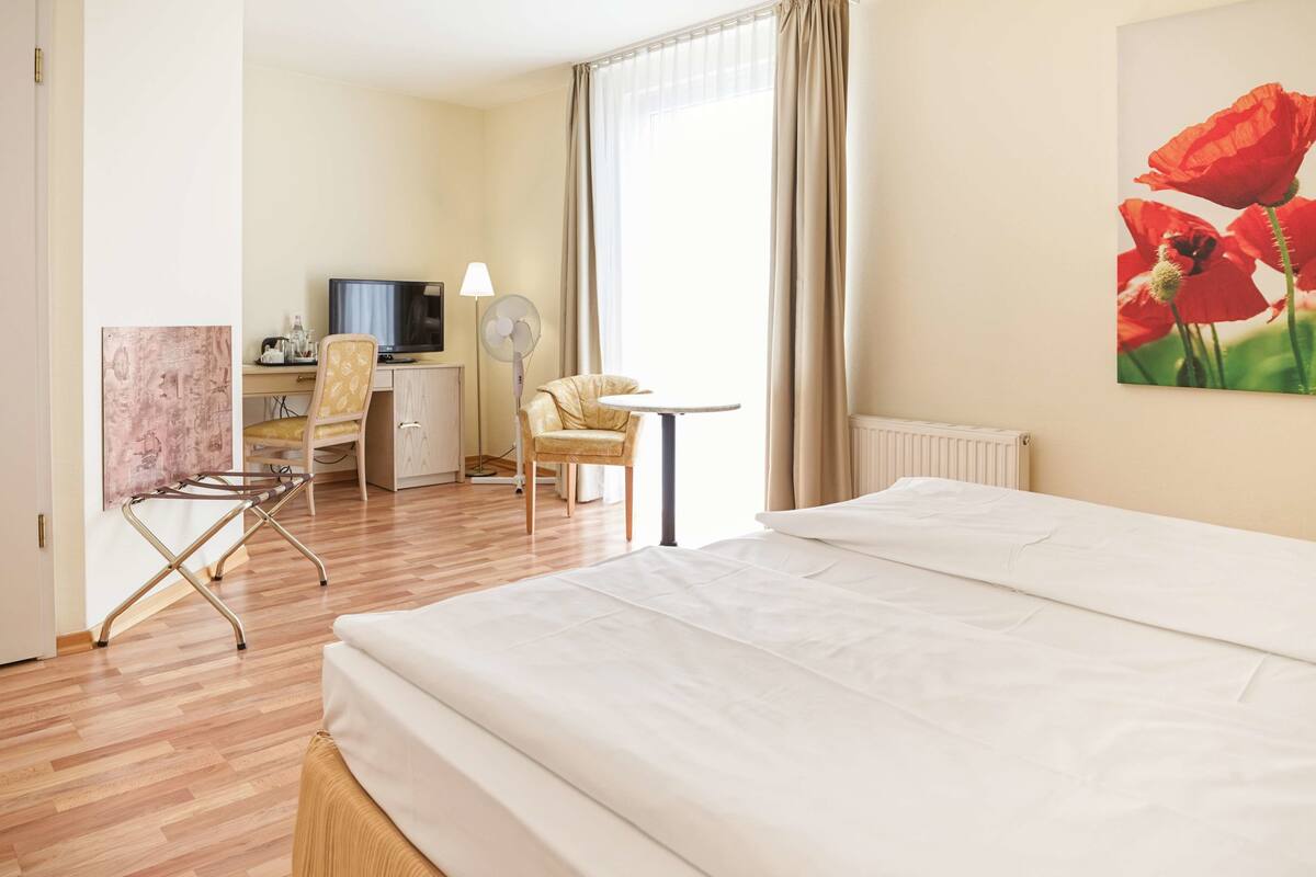 Ein Hotelzimmer mit einem Bett, einem Schreibtisch mit Computer, einem Sessel und einer Lampe, sowie einem Gemälde mit roten Blumen.