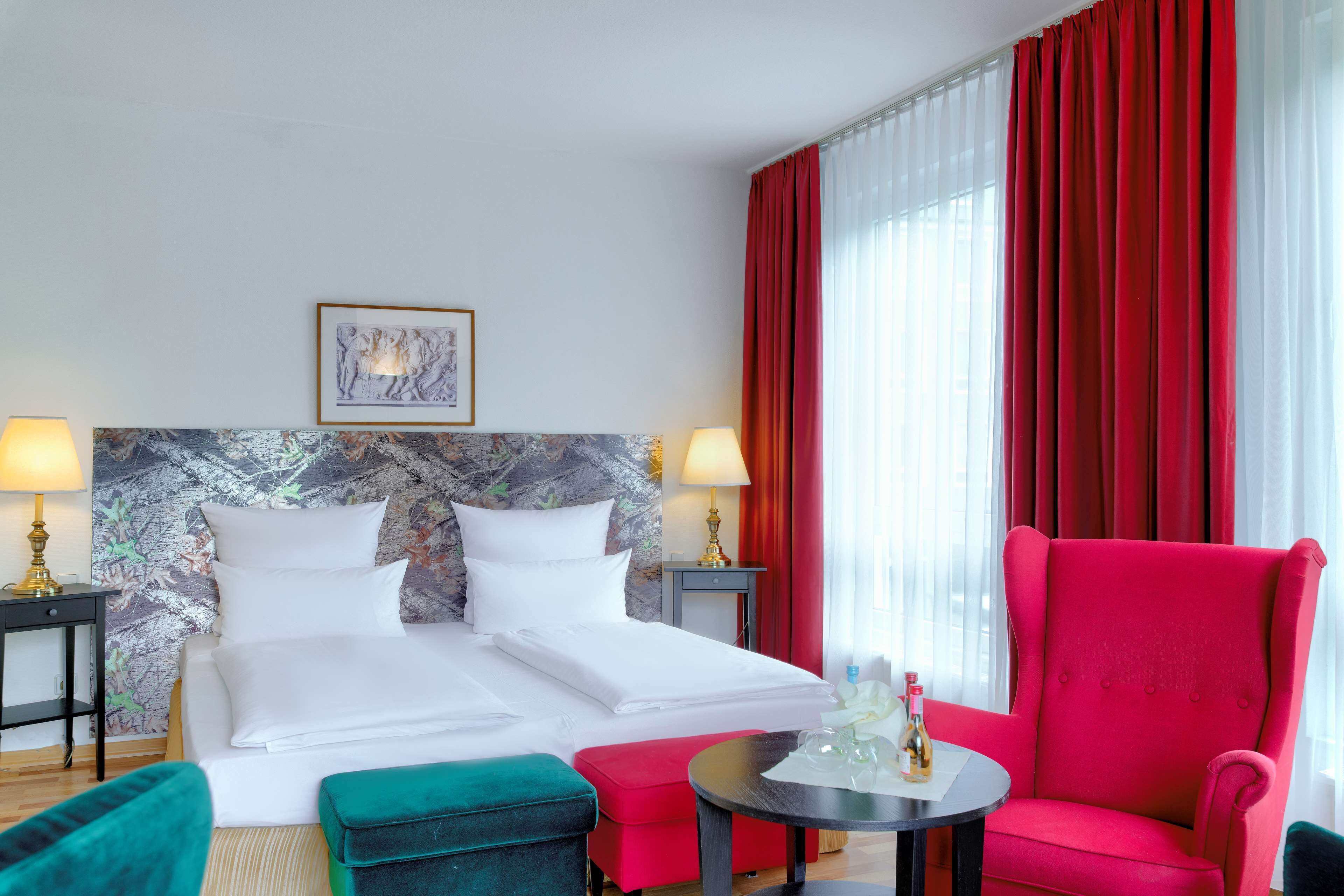 A hotel room with a bed, two bedside lamps, a red armchair, a small table, and a red ottoman.
