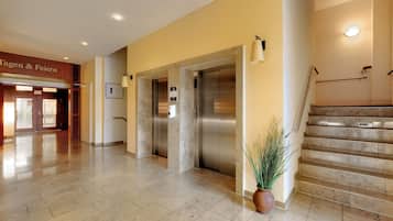 A hotel lobby with a reception desk, two elevators, a staircase, and a potted plant.