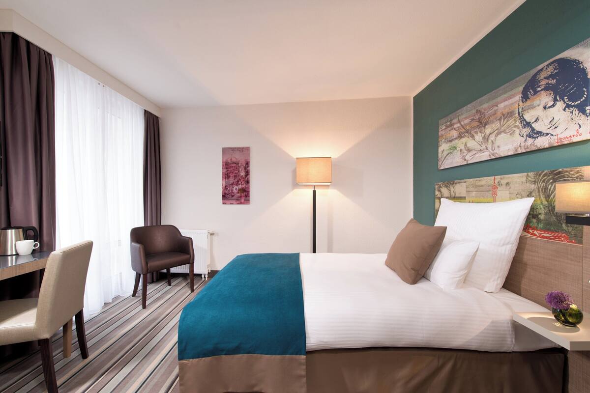 A hotel room with a bed, a desk, a chair, a lamp, and artwork on the walls.