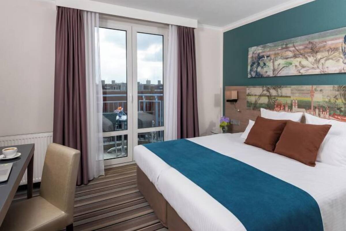 A hotel room with a bed, a desk, a chair, a window with curtains, and a painting on the wall.