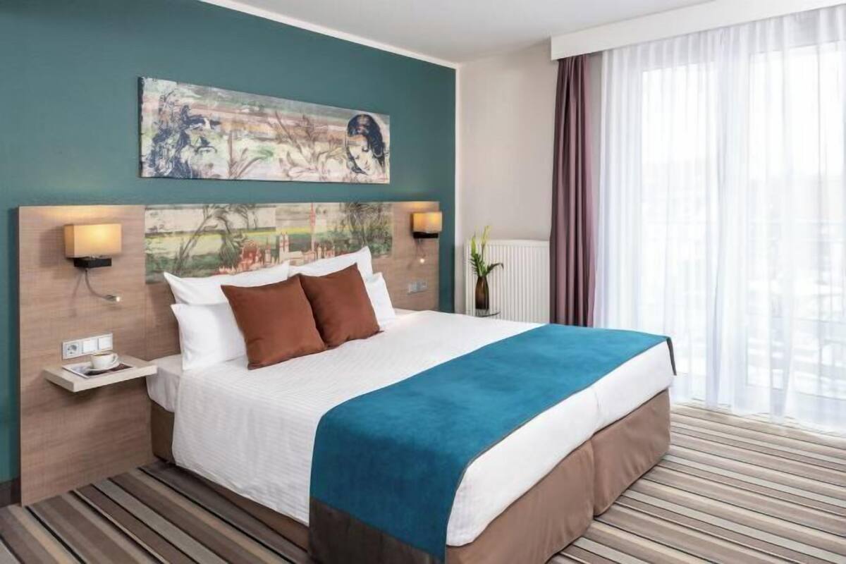 A hotel room with a large bed, two bedside tables with lamps, a painting above the bed, and a window with curtains.