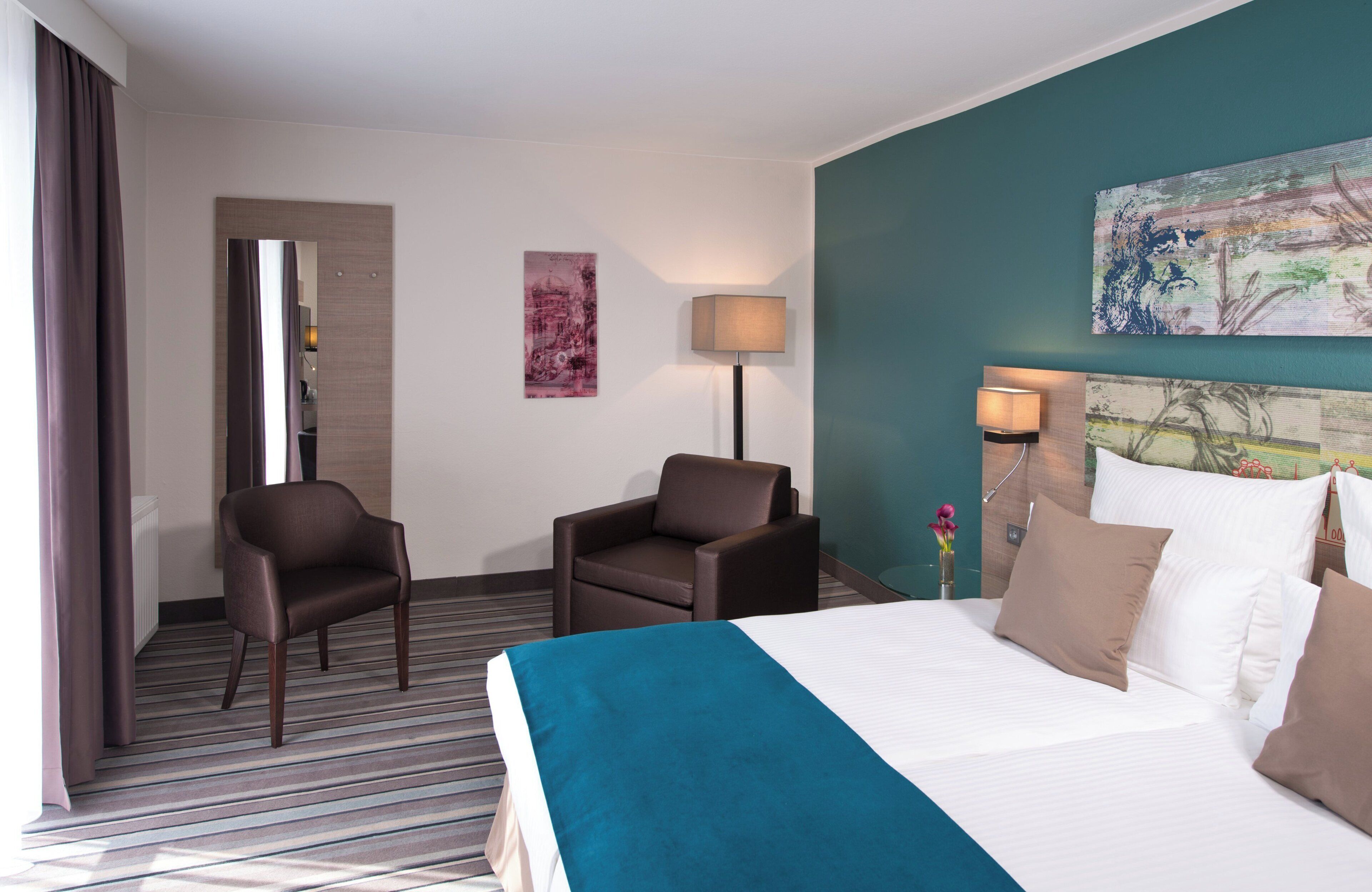 Ein Hotelzimmer mit einem Bett, zwei Stühlen, einem Schreibtisch, einer Lampe und Bildern an den Wänden.