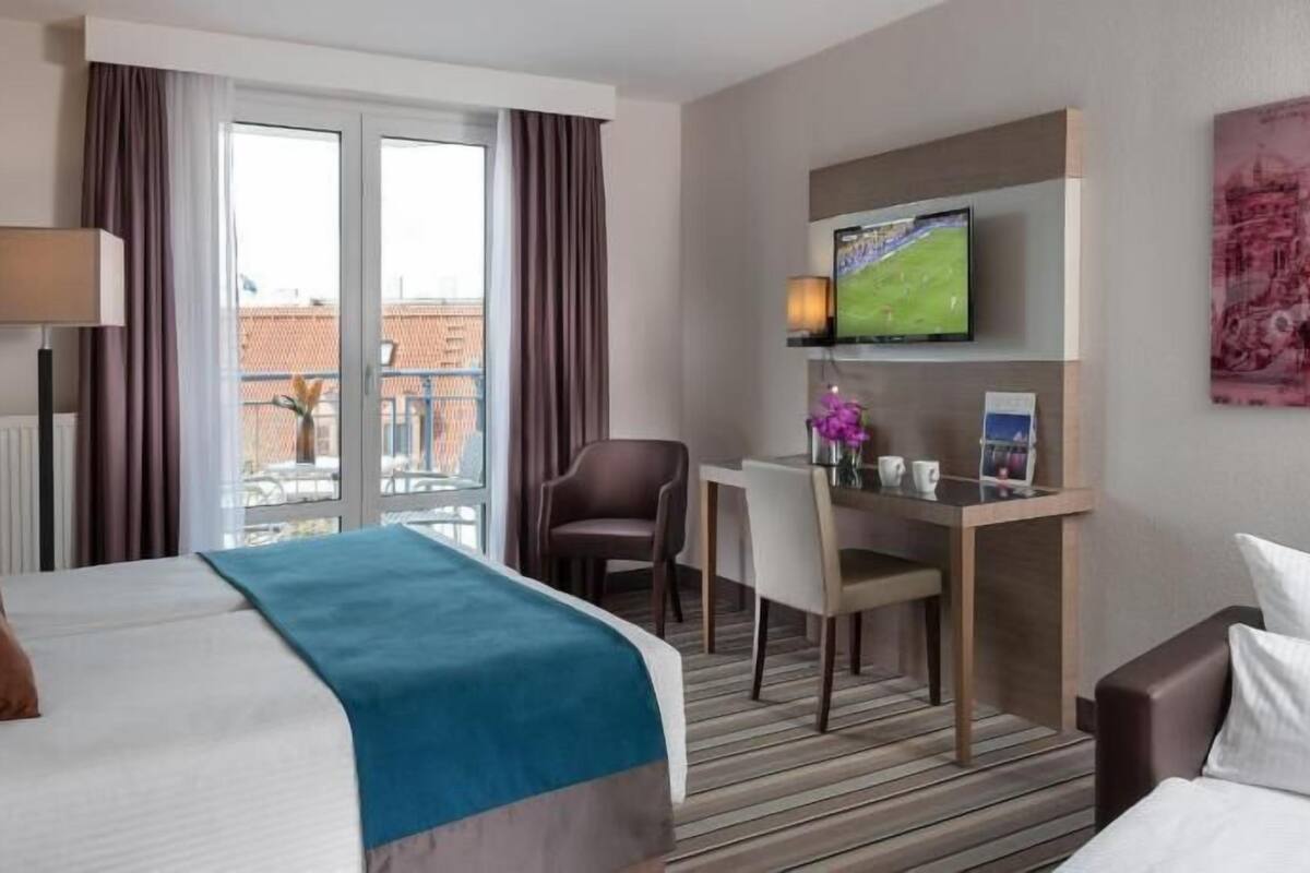 A hotel room with a bed, a desk with a chair, a television, and a window with curtains.