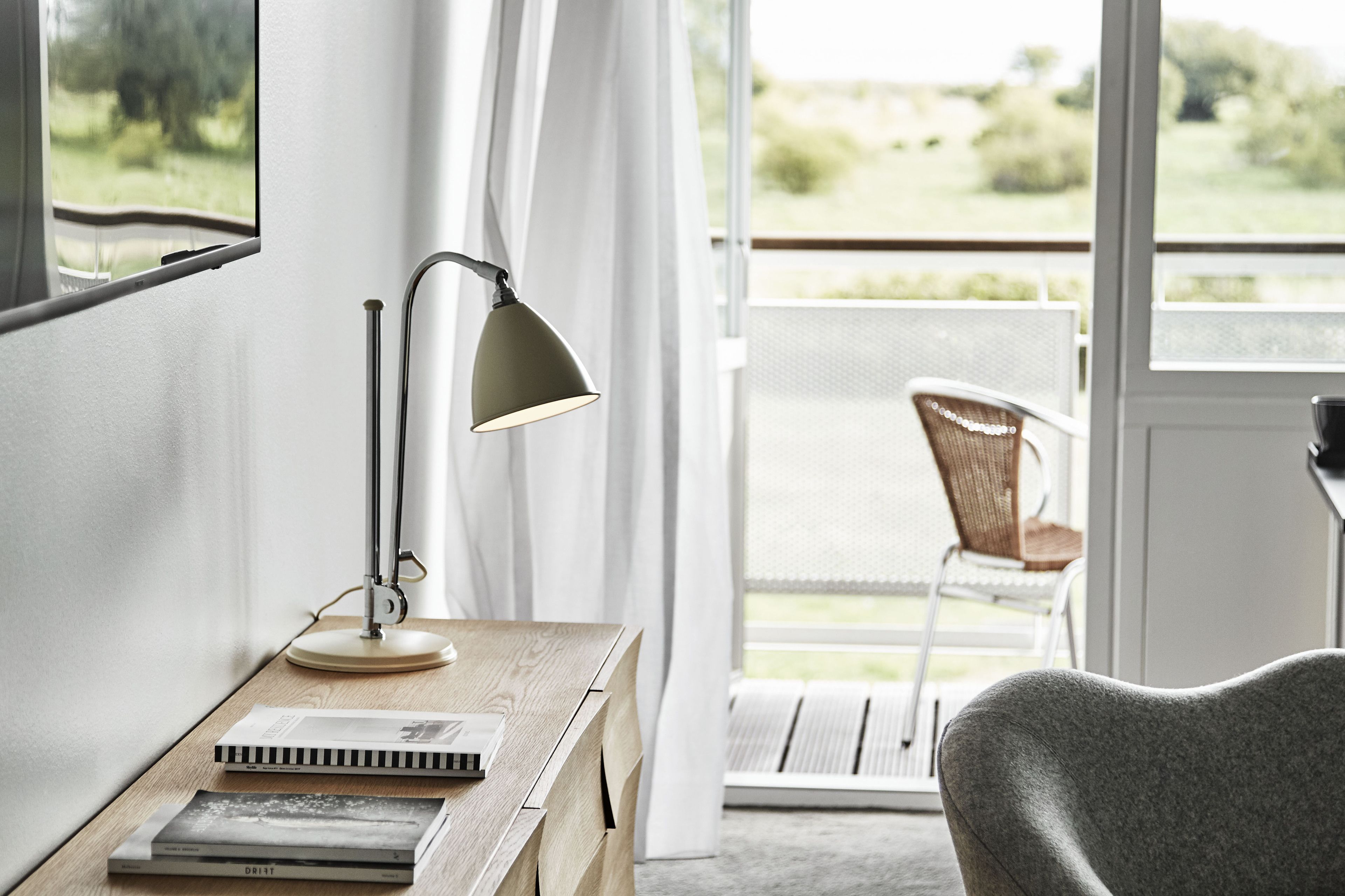 A room with a wooden desk, a lamp, a chair, and a view of a balcony with a chair and greenery outside.
