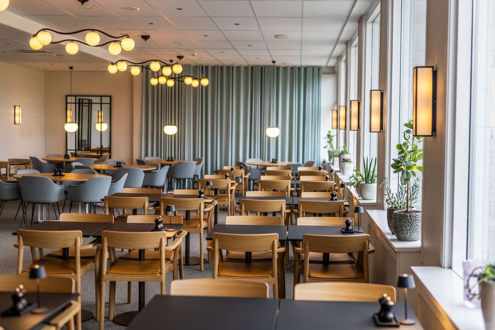 A hotel dining area with multiple tables and chairs, pendant lighting, and large windows with curtains.