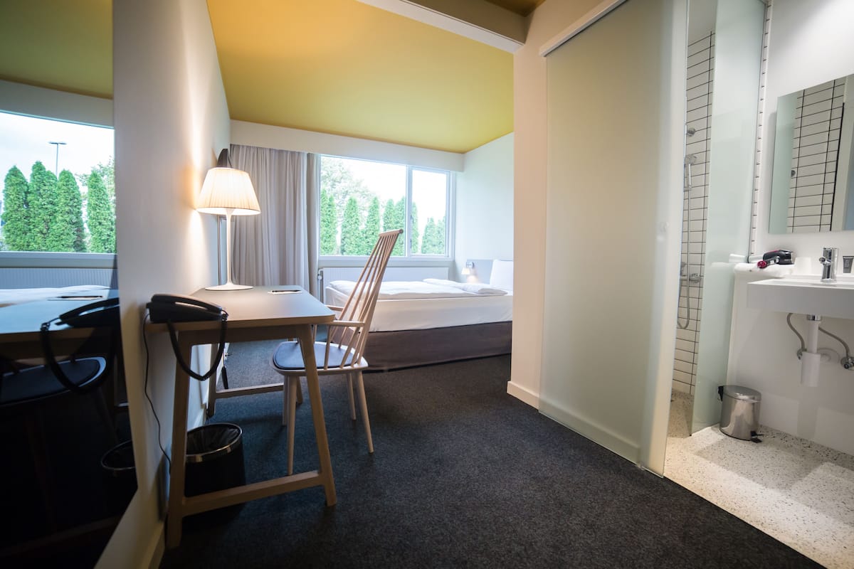 Ein Hotelzimmer mit Schreibtisch, Stuhl, Lampe und einem großen Fenster mit Blick auf Bäume.