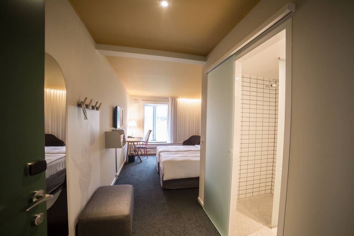 Ein Hotelzimmer mit zwei Betten, einem Schreibtisch, einem Stuhl, einer Lampe und einem Badezimmer mit Dusche.