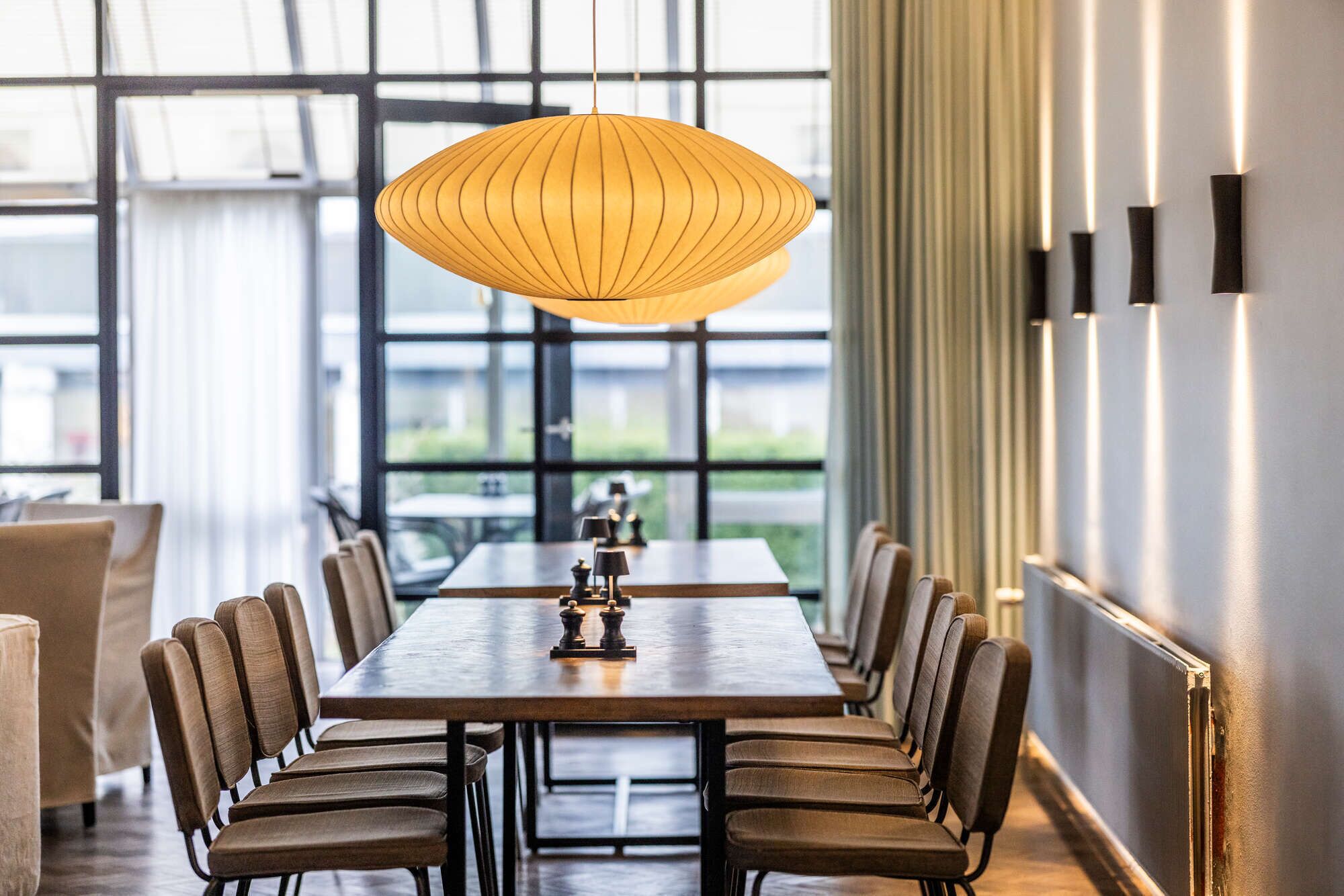 A dining area with a long table, multiple chairs, a hanging pendant light, and a view through large windows.