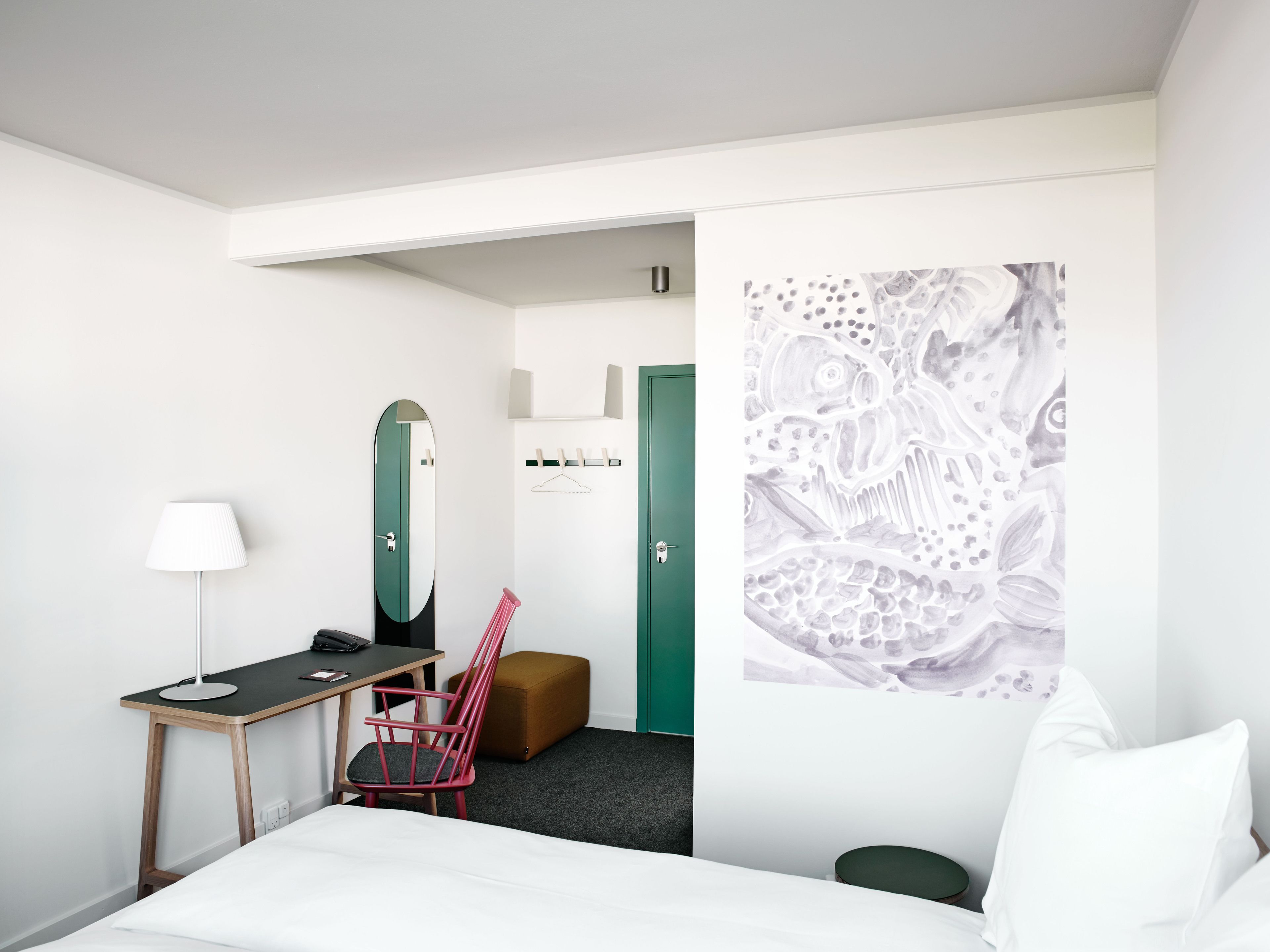 A hotel room with a bed, a desk with a lamp, a chair, a green door, and a wall with a large abstract painting.