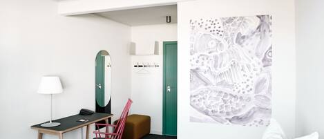 A hotel room with a bed, a desk with a lamp, a chair, a green door, and a wall with a large abstract painting.