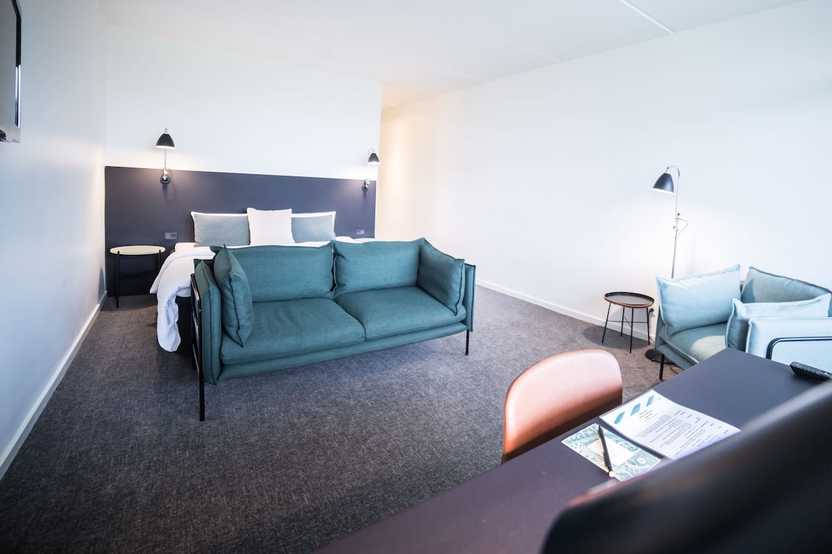 Ein modernes Hotelzimmer mit einem türkisfarbenen Sofa, einem kleinen runden Tisch, einem Schreibtisch mit Stuhl und einem Bett mit Kopfteil.