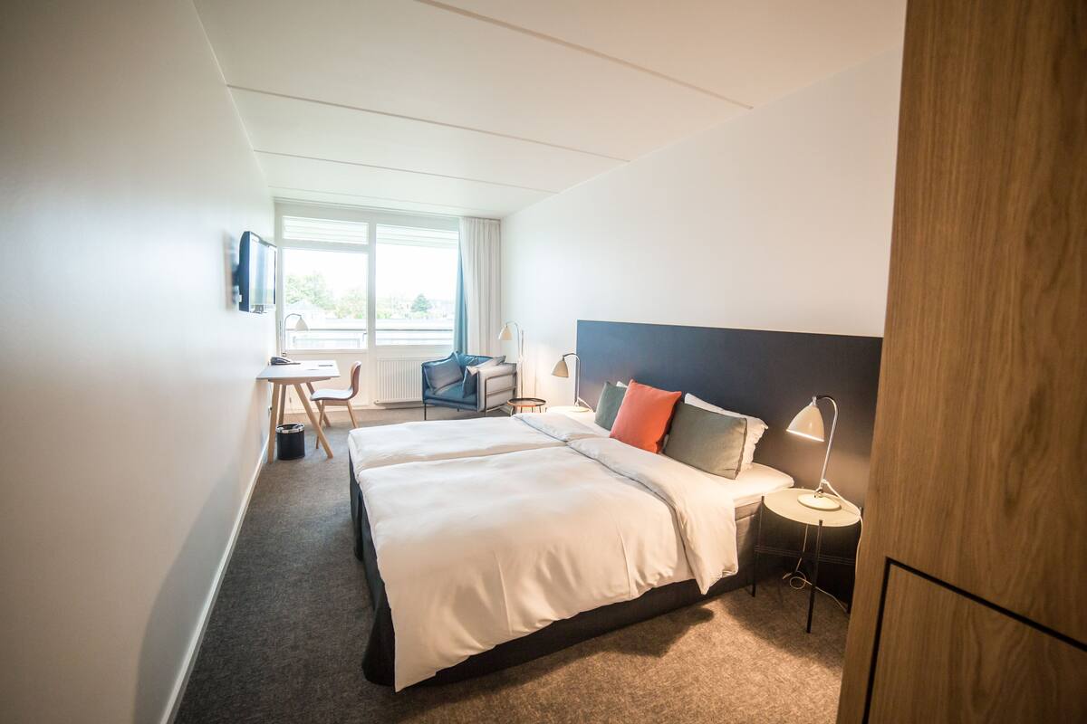 Ein Hotelzimmer mit einem großen Bett, einem Schreibtisch mit Stuhl, einem kleinen Tisch und einer Lampe.