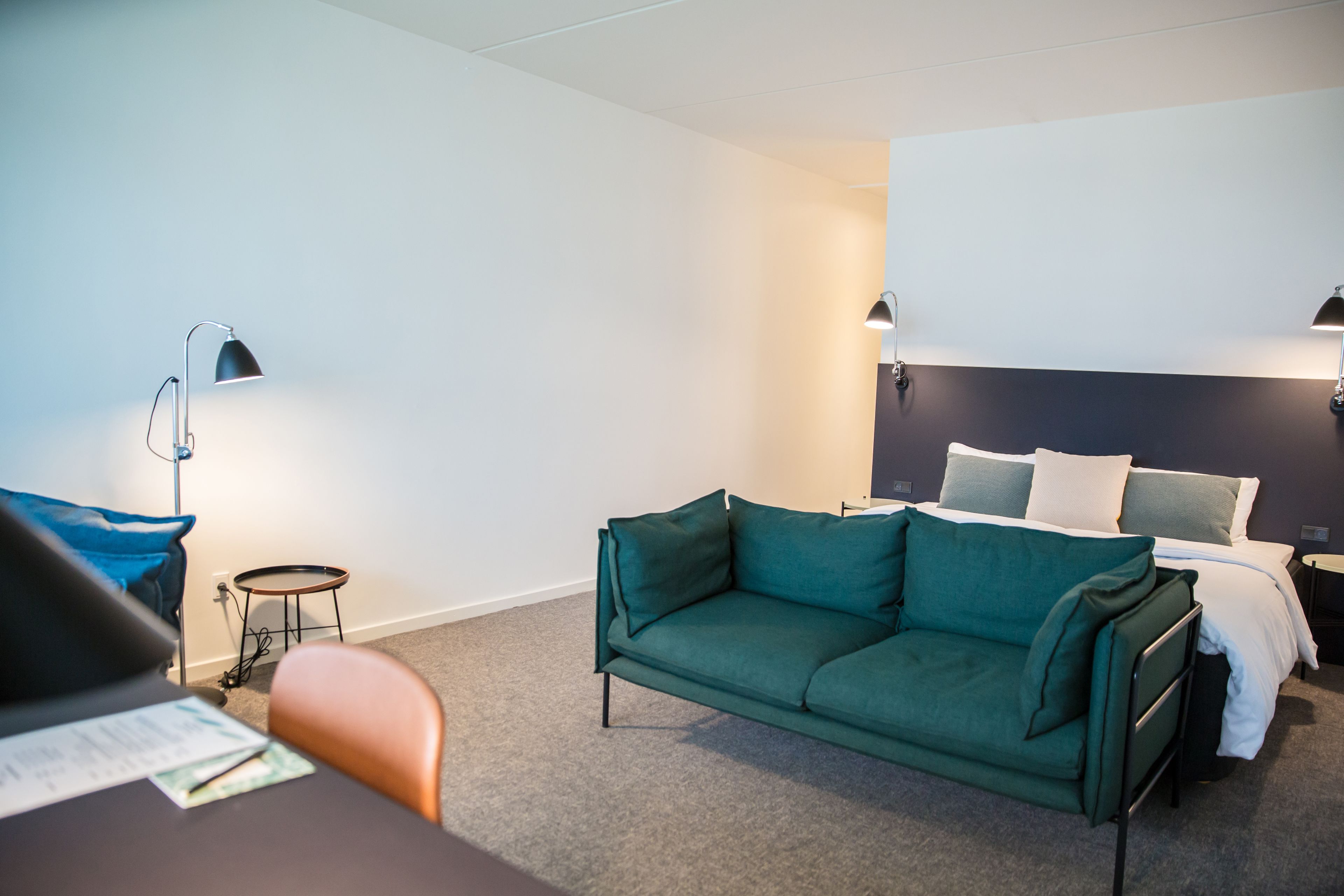 A hotel room with a green sofa, a small round table, a desk with papers, a bed with white bedding, and two wall-mounted lamps.