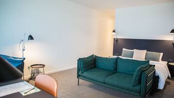 A hotel room with a green sofa, a small round table, a desk with papers, a bed with white bedding, and two wall-mounted lamps.