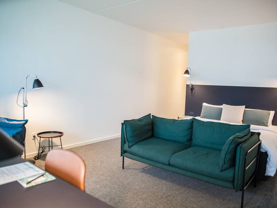 A hotel room with a green sofa, a small round table, a desk with papers, a bed with white bedding, and two wall-mounted lamps.