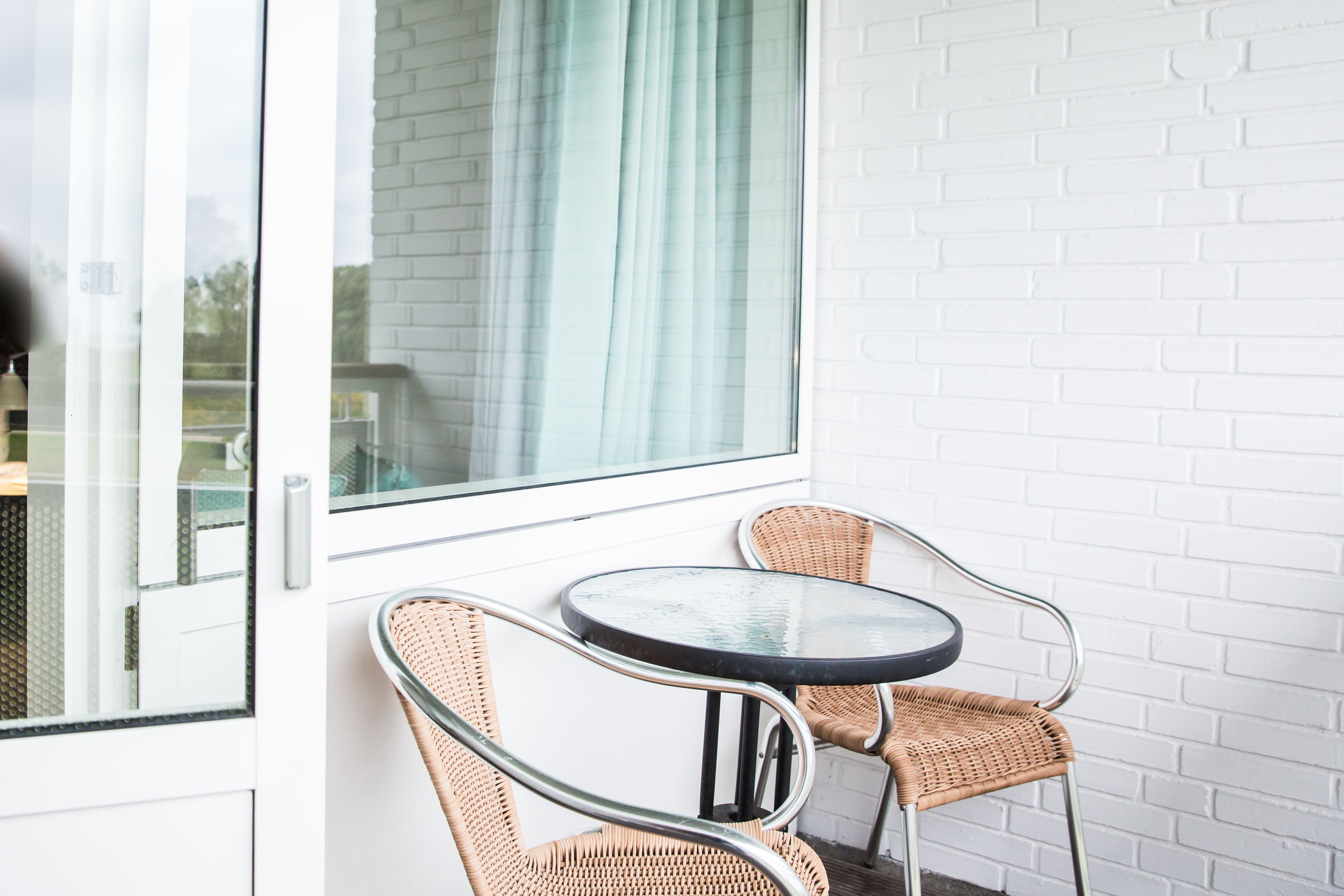 A small, round table with two wicker chairs, positioned next to a white brick wall and a window with a curtain.