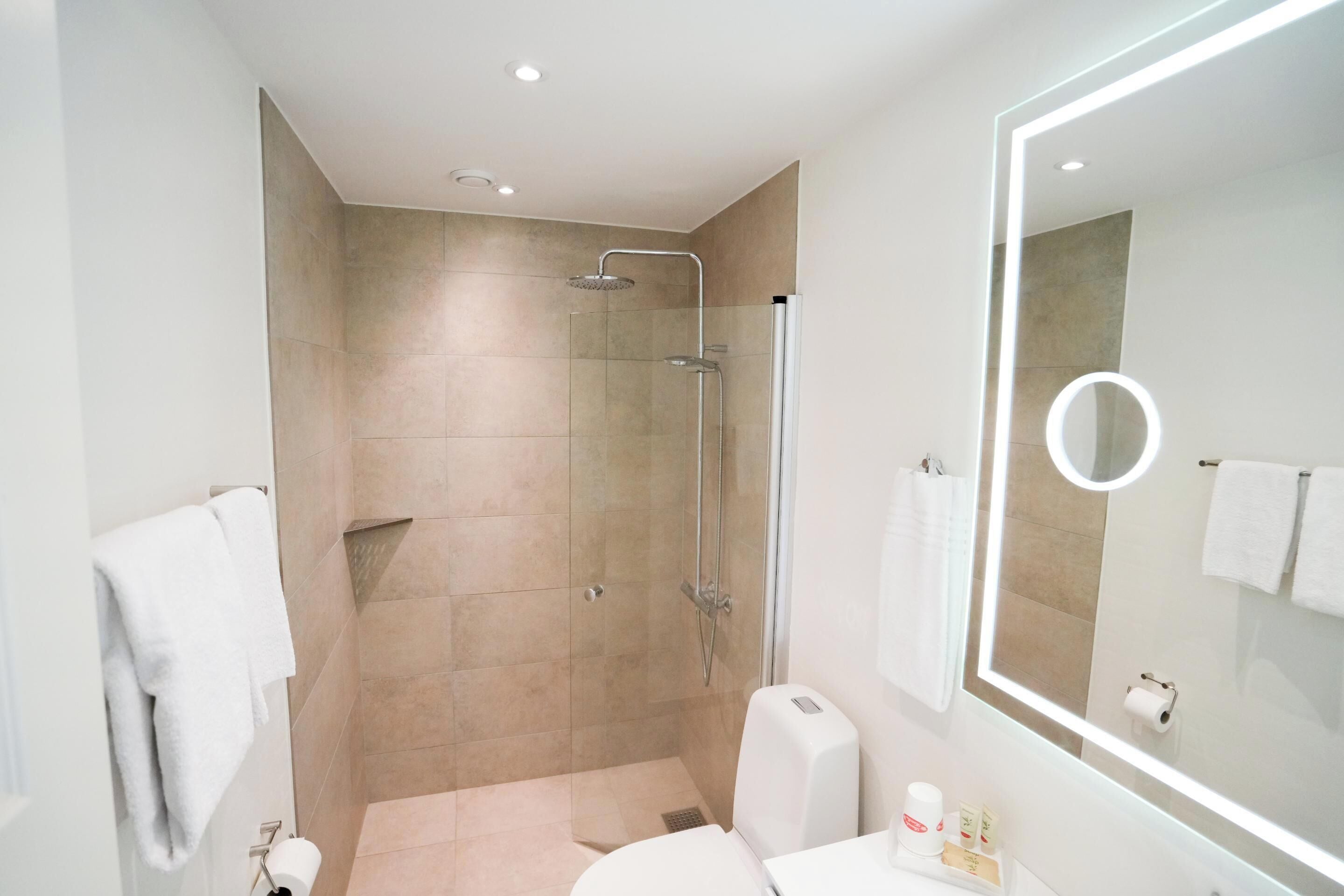A modern bathroom with a glass-enclosed shower, a large mirror with a lighted frame, and a white sink with toiletries.
