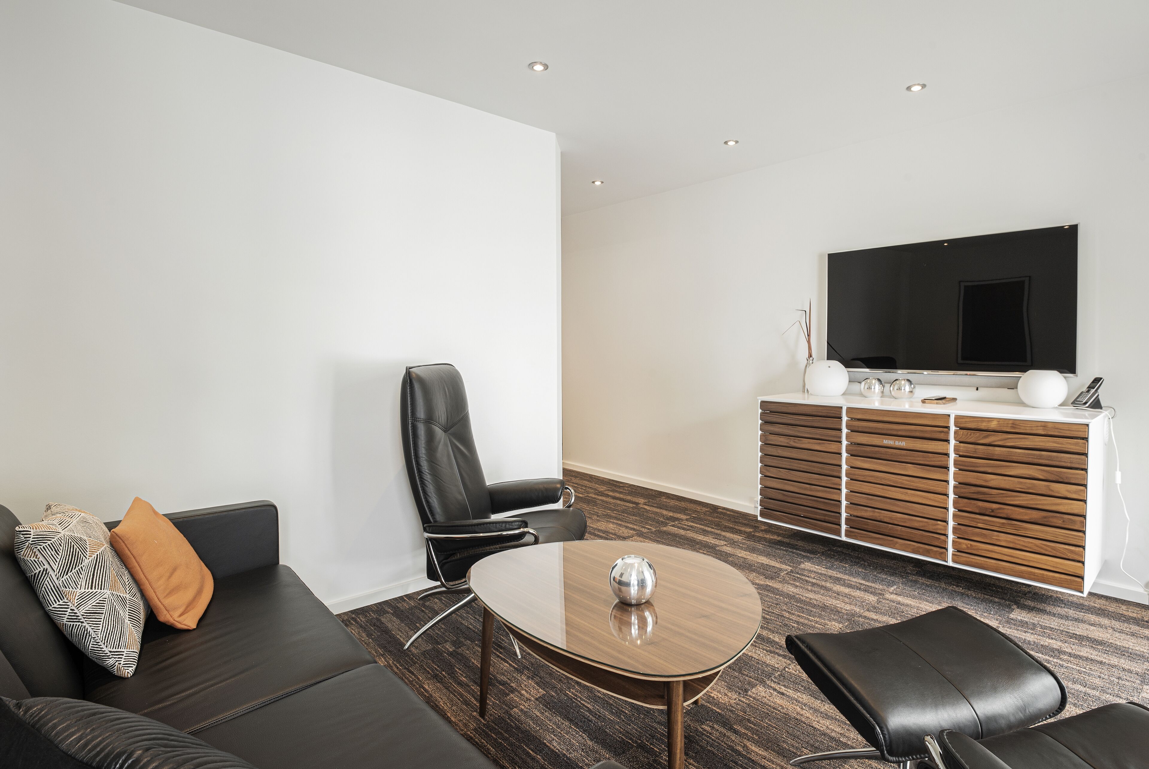 A modern living room with a black leather sofa, a round coffee table, and a black leather chair. A large flat-screen TV is mounted on the wall above a wooden cabinet.