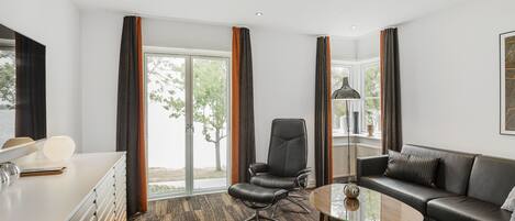 Una habitación de hotel moderna con un ventanal grande, un sofá de cuero, un escritorio con lámpara y una mesa de centro.