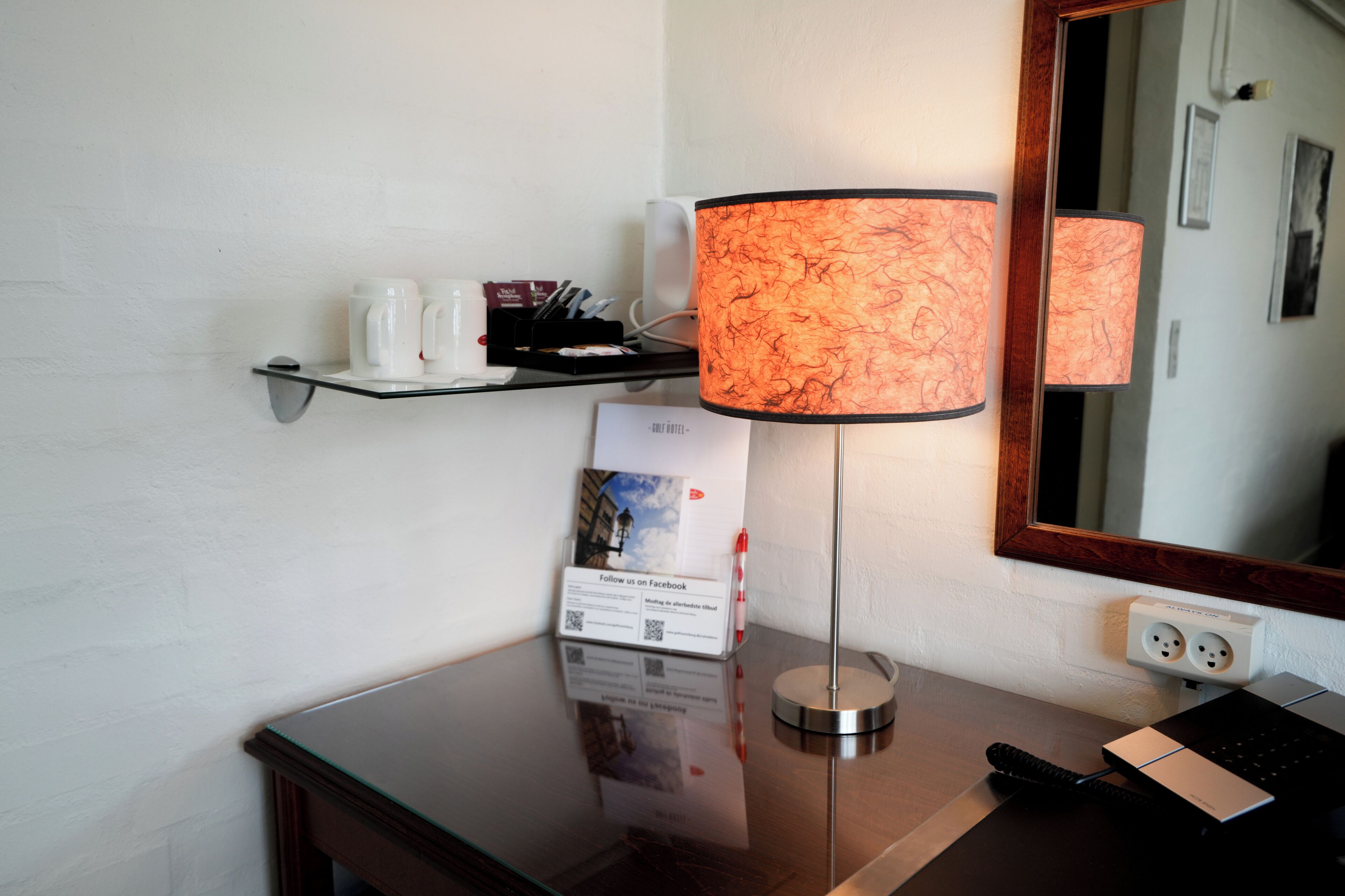 Habitación de hotel con una mesa de vidrio, una lámpara, un espejo y una estantería con una cafetera y un cartel.