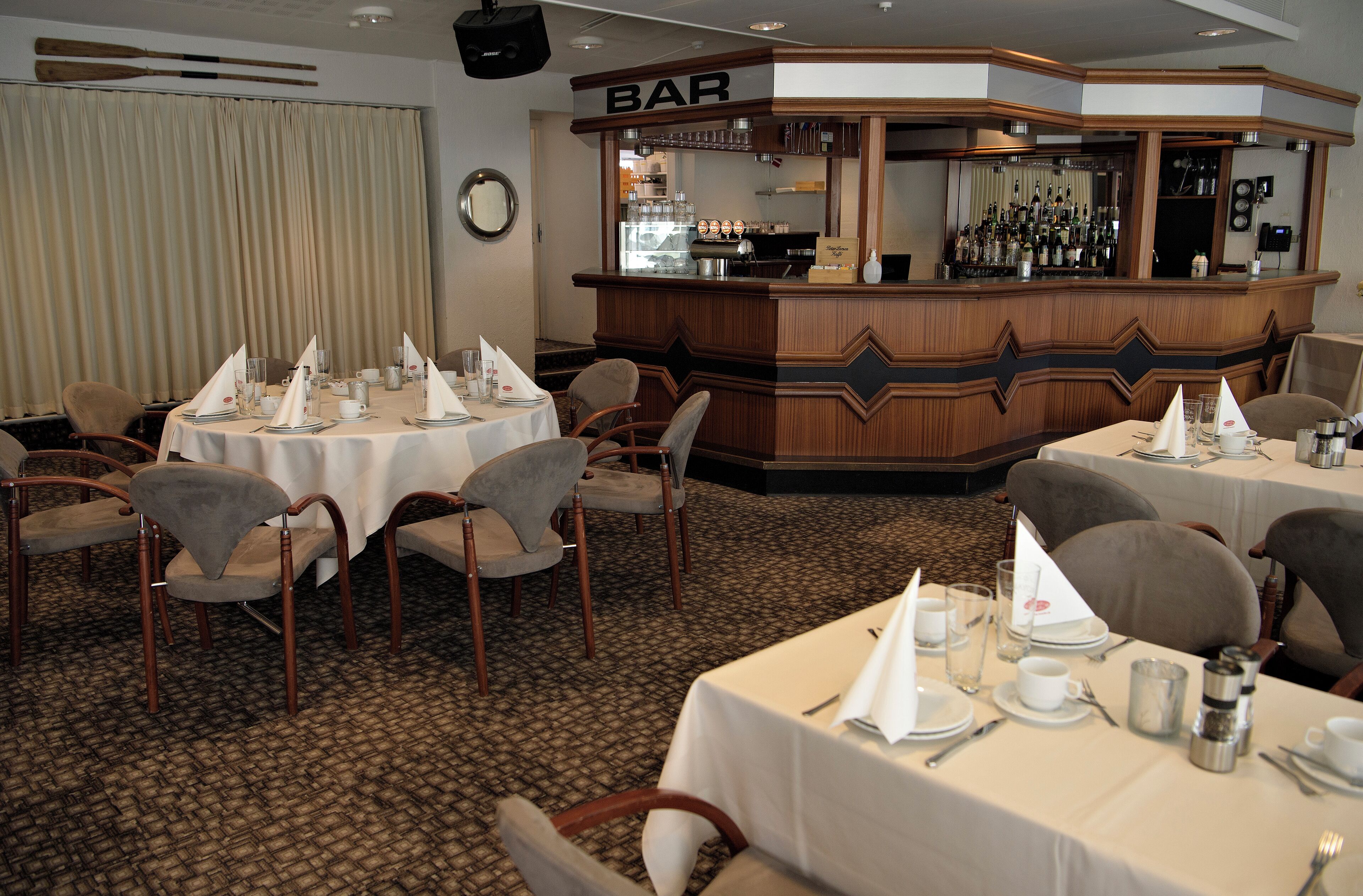 A restaurant with round tables set for dining, a bar area with a wooden counter, and a carpeted floor.