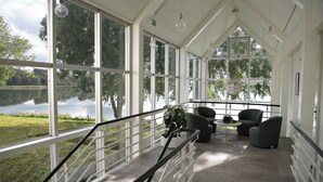 A spacious room with large windows overlooking a lake, a staircase with a glass railing, and modern furniture. - Golf Hotel Viborg (Viborg)